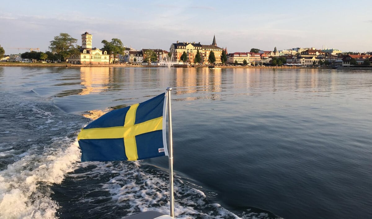 Svensk flagga bak på en båt, vatten och en stadslinje i bakgrunden